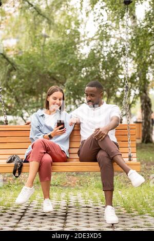 Jeune femme brune en chic décontracté montrant son petit ami africain une vidéo curieuse dans votre smartphone pendant que vous êtes assis sur votre swing Banque D'Images