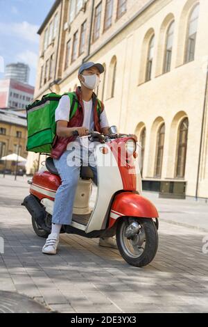 Livraison en toute sécurité. Plan vertical d'un jeune homme de messagerie avec sac thermo portant un masque de protection du visage livrant des aliments du restaurant par scooter automobile pendant l'épidémie de coronavirus Banque D'Images