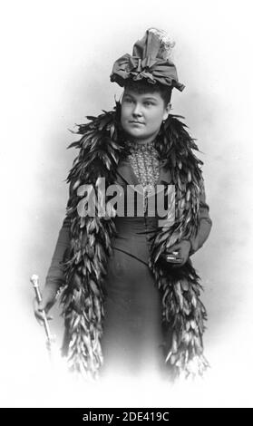 Vers 1890, cette jeune femme a visité son studio de photographie local pour faire une image permanente d'elle-même portant son nouveau boa plume., qui a atteint ses genoux. Elle porte également un beau chapeau de rubans enveloppés de façon extravagante, surmonté de plumes blanches. Elle semble ne porter aucun maquillage. Dans sa main droite se trouve une canne à tête argentée. Elle porte des gants sombres, et dans sa main gauche se trouve un petit compact, ou un porte-cigarette, ou ??? Pour voir mes autres images dignes de mention de femmes intéressantes, recherchez: Femme d'époque de pressor Banque D'Images