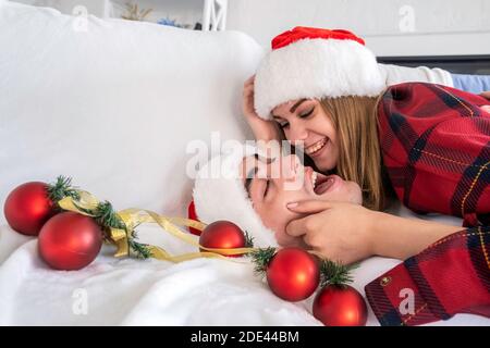 Jeune femme gaie se trouve sur l'homme portant le chapeau de santa et le pyjama. Couple sourit et apprécie l'ambiance de Noël entouré de boules d'arbre de Noël Banque D'Images