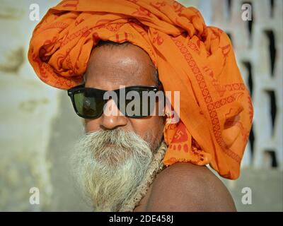 Le vieux hindou indien à barbe sadhu baba protège sa tête contre les rayons du soleil avec un foulard à tête émiettée orange et porte des lunettes de soleil sombres. Banque D'Images