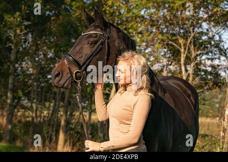 Vue avant, portrait. Femme dans les stands de vêtements décontractés dans la forêt avec son cheval. Dans le soleil d'automne. Banque D'Images
