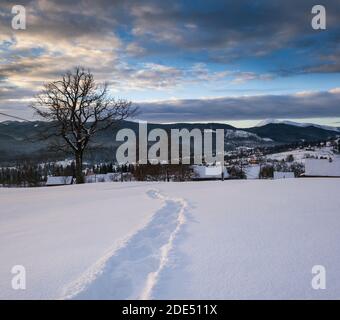 Petit village alpin et hiver montagne enneigée au premier lever du soleil autour, Voronenko, Carpates, Ukraine. Tracer sur un chemin fraîchement trodden à travers g Banque D'Images