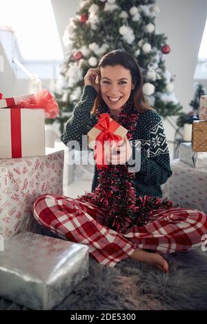 Belle brune caucasienne est sur le point d'ouvrir le cadeau de Noël, assis sur le sol dans la salle de séjour, souriant, ajuster les cheveux, regardant la caméra. Oeil Banque D'Images