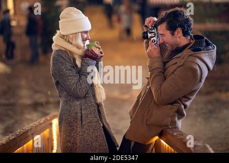 jeune homme caucasien prenant une photo de sa petite amie tenant une tasse de thé à l'extérieur avec l'ancien appareil photo. concept de christmastime Banque D'Images