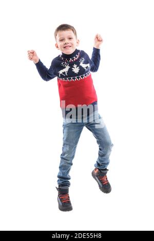 Un petit garçon excités qui fête et saute avec des mains levées regardant l'appareil photo dans le pull de Noël. Portrait du corps entier isolé sur fond blanc. Banque D'Images