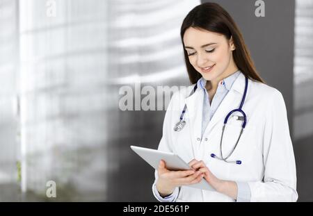 Gaie et intelligente femme-médecin tient une tablette dans ses mains, alors qu'elle est debout dans une clinique. Portrait d'une femme médecin sympathique Banque D'Images