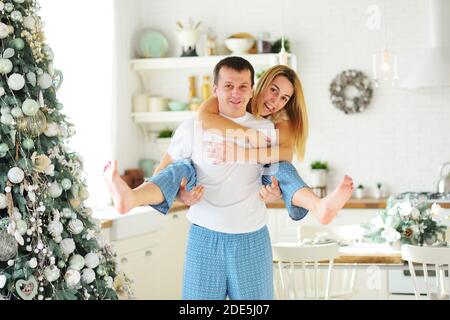 Jeune couple - un homme et une femme en pyjama embrassant et se berner sur le fond d'une cuisine moderne et d'un arbre de Noël. Banque D'Images