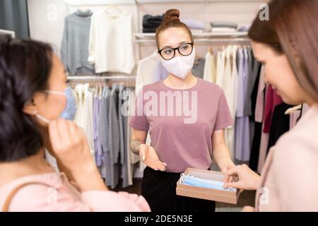 Jeune assistante de boutique contemporaine dans les lunettes recommandant des femmes visiteurs à prendre des masques de protection avant d'entrer dans le rayon Banque D'Images