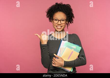 Femme enseignante ou tutrice afro-américaine isolée sur un mur rose de studio. Une fille étudiante porte des lunettes tenant des carnets, souriant, montrant le doigt sur l'espace libre. Banque D'Images