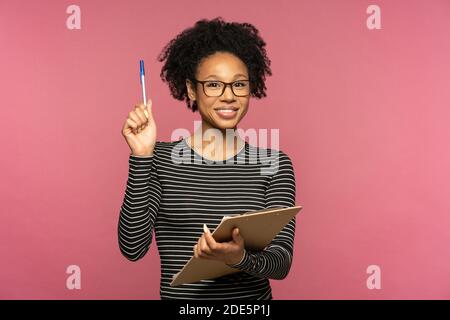 Jeune femme enseignante afro-américaine heureuse isolée sur le mur rose du studio. Une fille étudiante porte des lunettes tenant une chemise et un stylo, souriant, regardant l'appareil photo. Ed Banque D'Images