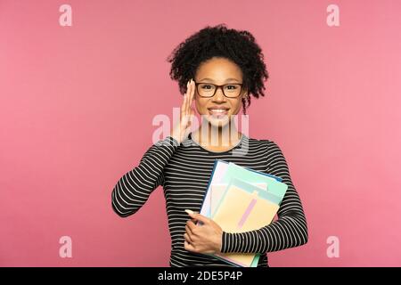 Jeune femme enseignante ou enseignante afro-américaine heureuse isolée sur un mur rose de studio. Une fille étudiante porte des lunettes tenant des carnets et souriant. L'éducation en h Banque D'Images