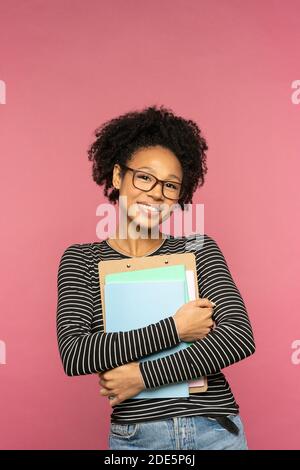 Jeune femme enseignante ou enseignante afro-américaine heureuse isolée sur un mur rose de studio. Une fille étudiante porte des lunettes tenant des carnets et souriant. Collège et Bonjour Banque D'Images