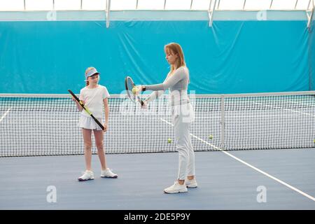 Adorable fille adolescente en vêtements de sport regardant la femme entraîneur et à l'écoute de ses instructions sur la façon d'utiliser le ballon et racket pendant le jeu Banque D'Images