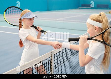 Deux filles adolescentes heureuses en vêtements d'activité tenant des raquettes de tennis pendant serrer les mains par-dessus le filet contre le terrain avant la nouvelle formation stade moderne Banque D'Images