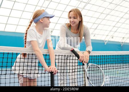 Jolie fille blonde en vêtements de sport blancs et son entraîneur avec raquettes de tennis contre le filet au stade devant caméra et interaction Banque D'Images