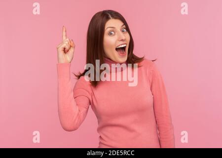 Une femme excitée et inspirée avec des cheveux bruns pointant vers le haut en regardant l'appareil photo avec un sourire crasseux, heureuse avec une nouvelle idée géniale, trouver la solution. Studio intérieur Banque D'Images
