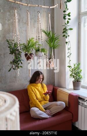 Femme indépendante en chandail jaune, reposant, assis sur un canapé sous un cintre en coton macrame plante avec des plantes à la maison, en utilisant le téléphone mobile. Passe-temps, fait à la main, Banque D'Images