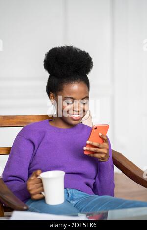 Gros plan d'Afro-américaine femme millénaire porter un chandail violet assis dans un fauteuil, en utilisant un smartphone mobile, en discutant dans les réseaux sociaux, en buvant te Banque D'Images