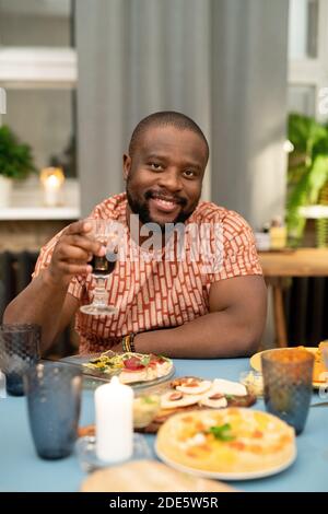 Joyeux jeune Africain qui applaudisse avec un verre de rouge vin devant l'appareil photo pendant que vous êtes assis près de l'hôtel, servi de fête Table pendant le dîner de Noël Banque D'Images