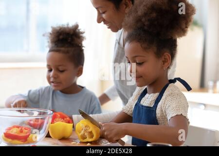 Une mère d'accueil noire souriante regarde les enfants adoptés cuisiner des plats de légumes Banque D'Images