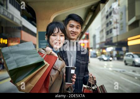 un jeune couple asiatique heureux attend le bus dans une ville moderne avec des sacs à provisions à portée de main Banque D'Images