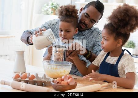 Le père africain attentionné enseignant aux enfants frères et sœurs soupire la farine pour le gâteau Banque D'Images