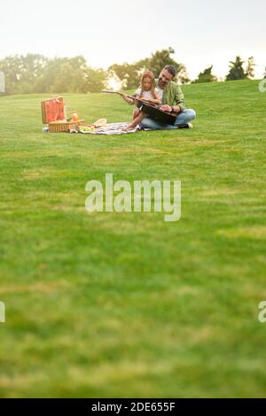 Photo pleine longueur d'une petite fille heureuse assise avec elle père aimant sur une herbe verte dans le parc et l'apprentissage comment jouer de la guitare Banque D'Images