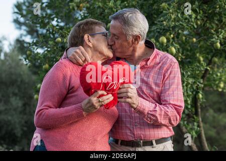 Couple senior romantique avec cœur rouge embrassant et embrassant doucement tout en célébrant l'anniversaire de mariage Banque D'Images