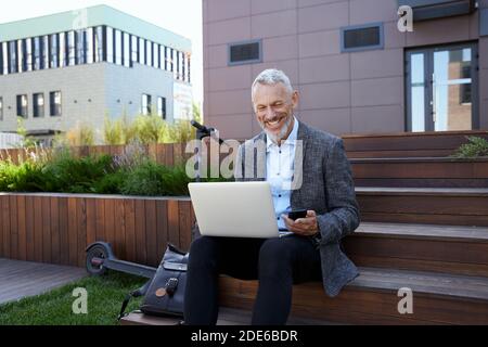 Restez en contact n'importe où. Homme d'affaires mûr souriant, passant un appel vidéo tout en travaillant sur un ordinateur portable, assis à l'extérieur pendant la journée Banque D'Images