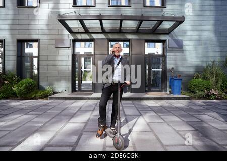 Photo en longueur d'un homme d'affaires mûr souriant, parlant au téléphone tout en se tenant debout avec un scooter électrique près d'un immeuble de bureau moderne Banque D'Images