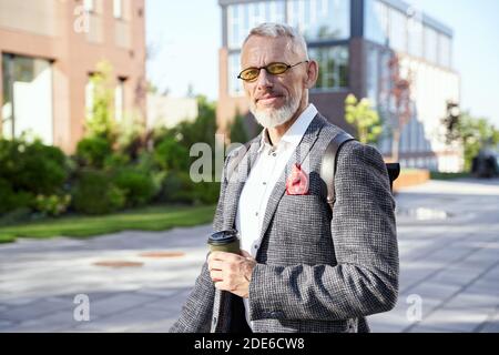 Homme occupé. Portrait d'un homme d'affaires d'âge moyen plein de confiance et élégant en lunettes de soleil regardant l'appareil photo tout en se tenant debout à l'extérieur avec une tasse de café Banque D'Images
