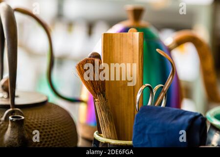 Un fouet à thé en bambou (chasen) et une paire de ciseaux se trouvent à côté d'un bloc de bois dans une tasse en céramique, prêts pour une cérémonie du thé japonaise au magasin de thé ANMO à Düsseldorf, en Allemagne. Une théière en fonte tetsubin se dresse devant une bouilloire laquée irisée multicolore en arrière-plan. Banque D'Images