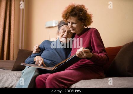 Femme âgée et sa fille adulte regardant un album photo ensemble sur un canapé dans le salon, parlant joyeusement de souvenirs. Week-end avec les parents, Banque D'Images