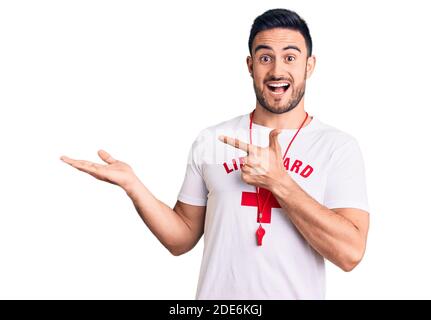 Jeune homme beau portant l'uniforme de maître-nageur stupéfié et souriant à l'appareil photo tout en présentant avec la main et pointant avec le doigt. Banque D'Images