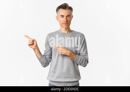 Portrait de l'homme gay adulte dans le chandail gris, pointant les doigts vers la gauche et montrant la publicité, debout sur fond blanc Banque D'Images