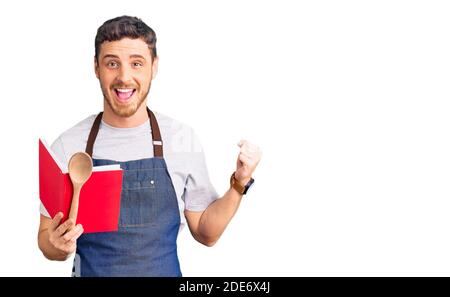 Beau jeune homme avec l'ours portant un tablier boulanger professionnel lecture livre de recettes de cuisine criant fier, célébrant la victoire et le succès très excite Banque D'Images