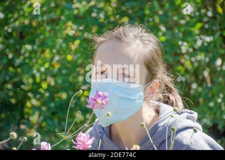 Jeune femme essayant de sentir des fleurs sauvages dans le parc tout en portant un masque médical. Photo conceptuelle du coronavirus ou de l'allergie, sortie de quarantaine. Banque D'Images