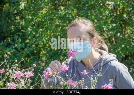 Jeune femme essayant de sentir des fleurs sauvages dans le parc tout en portant un masque médical. Photo conceptuelle du coronavirus ou de l'allergie, sortie de quarantaine. Banque D'Images