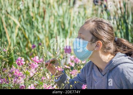 Jeune femme essayant de sentir des fleurs sauvages dans le parc tout en portant un masque médical. Photo conceptuelle du coronavirus ou de l'allergie, sortie de quarantaine. Banque D'Images