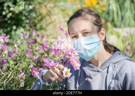 Jeune femme essayant de sentir des fleurs sauvages dans le parc tout en portant un masque médical. Photo conceptuelle du coronavirus ou de l'allergie, sortie de quarantaine. Banque D'Images