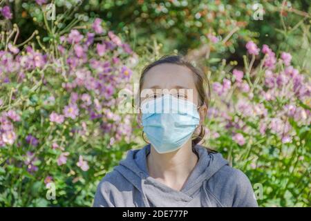 Jeune femme essayant de sentir des fleurs sauvages dans le parc tout en portant un masque médical. Photo conceptuelle du coronavirus ou de l'allergie, sortie de quarantaine. Banque D'Images