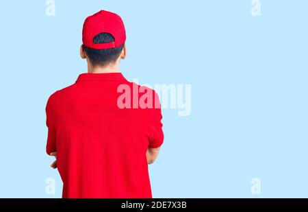 Jeune homme beau avec barbe portant l'uniforme de livraison debout en arrière regarder loin avec les bras croisés Banque D'Images