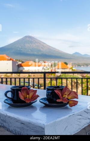 Two cups of coffee with flowers of Hibiscus Tiliaceus or Sea hibiscus, view of volcano Agung on the background. Amed, Karangasem, Bali, Indonesia. Stock Photo