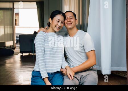 Portrait d'un jeune couple asiatique adulte qui s'embrasse avec l'intérieur de la maison en arrière-plan. 30s heureux mûr mari et femme souriant et regardant l'appareil photo. Concept de vie de mariage et de relation heureuse Banque D'Images