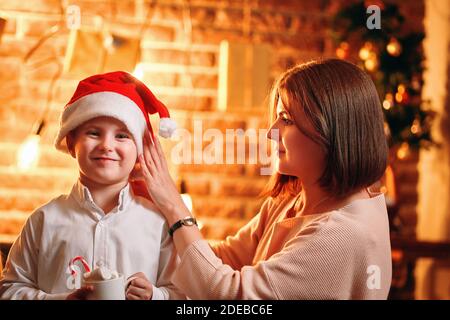 Belle mère met le chapeau du Père Noël sur son enfant. La famille de Noël. Tasse de chocolat chaud avec guimauves dans la main du garçon. Nouvel an. Boîtes-cadeaux, guirlandes et branches de sapin sur un mur en brique. Banque D'Images