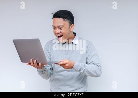 Wow visage de jeune asiatique homme a choqué ce qu'il voit dans l'ordinateur portable lors de travail isolé fond gris porter une chemise grise. Banque D'Images