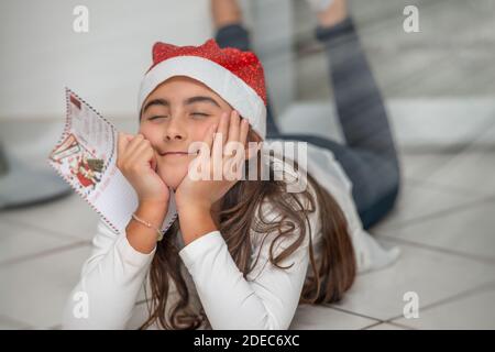 Une jeune fille couchée sur le sol à la maison portant un chapeau rouge de Noël tenant une lettre au Père Noël. Rêver de nouveaux jeux. Banque D'Images