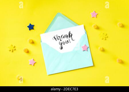 Carte de remerciement manuscrite dans une enveloppe bleu sarcelle, prise du haut sur fond jaune avec étoiles brillantes et bonbons Banque D'Images