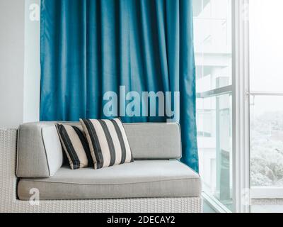 Confortable canapé-lit en rotin avec oreillers décorés sur le balcon et patio sur un bâtiment haut sur fond de rideau bleu près du verre porte à l'intérieur de la pièce Banque D'Images